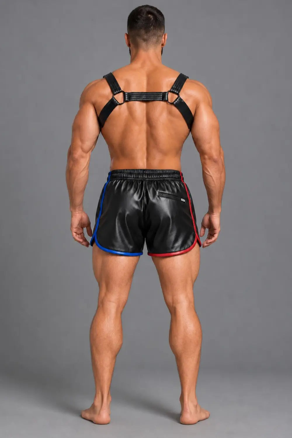 A muscular man viewed from the back wearing black leather athletic shorts with blue and red side piping and a black PU leather chest harness, photographed against a plain gray studio background